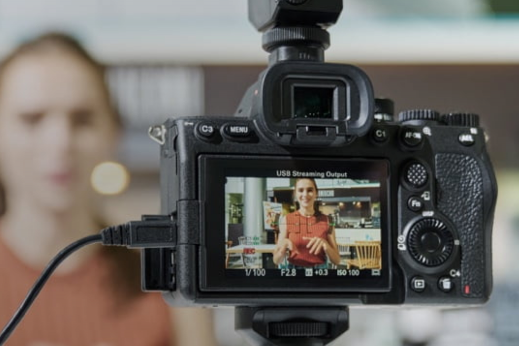 A Sony A7 IV filming a woman indoors, viewed from behind the camera with the rear LCD screen showing her speaking on video.
