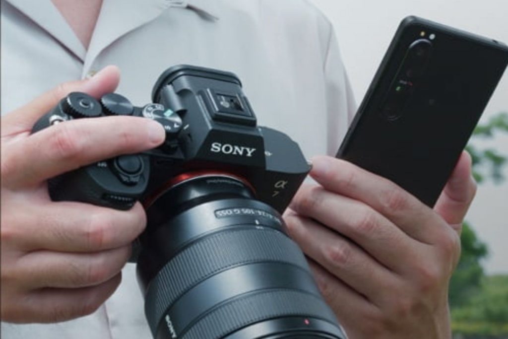 A person holding a Sony Alpha camera in one hand while checking settings or transferring files on a smartphone with the other.