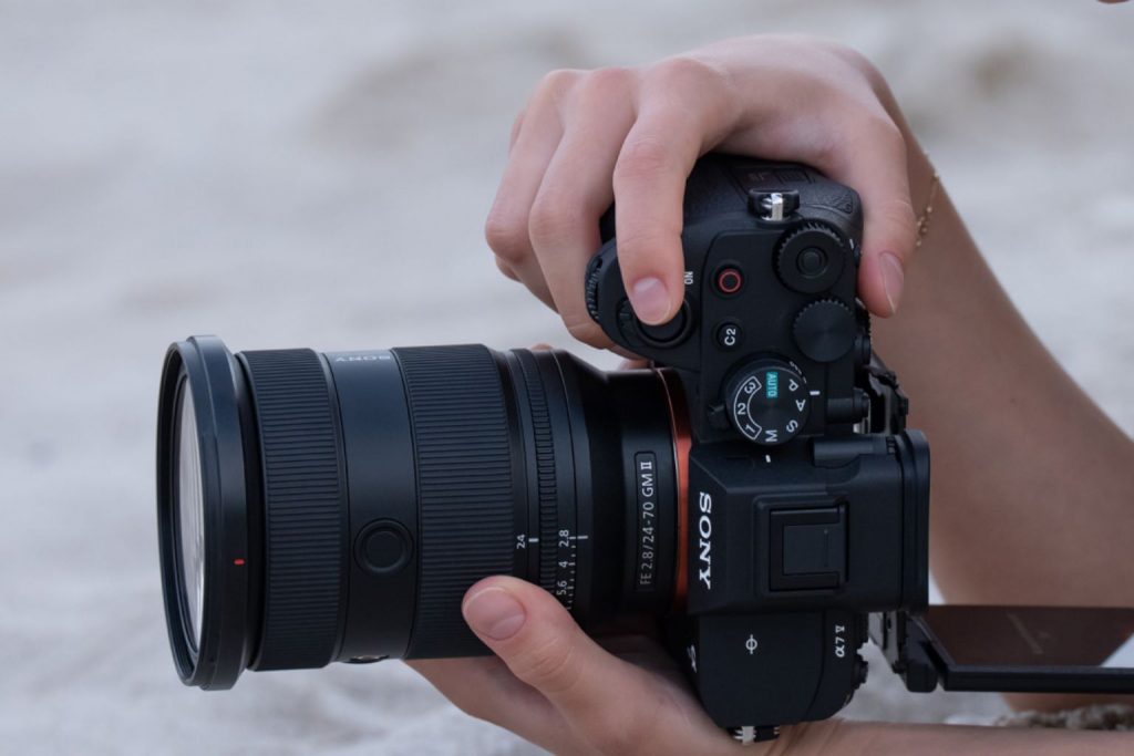 Close-up of hands holding the Sony A7 V camera with a 24–70mm G Master II lens, adjusting settings while shooting outdoors.