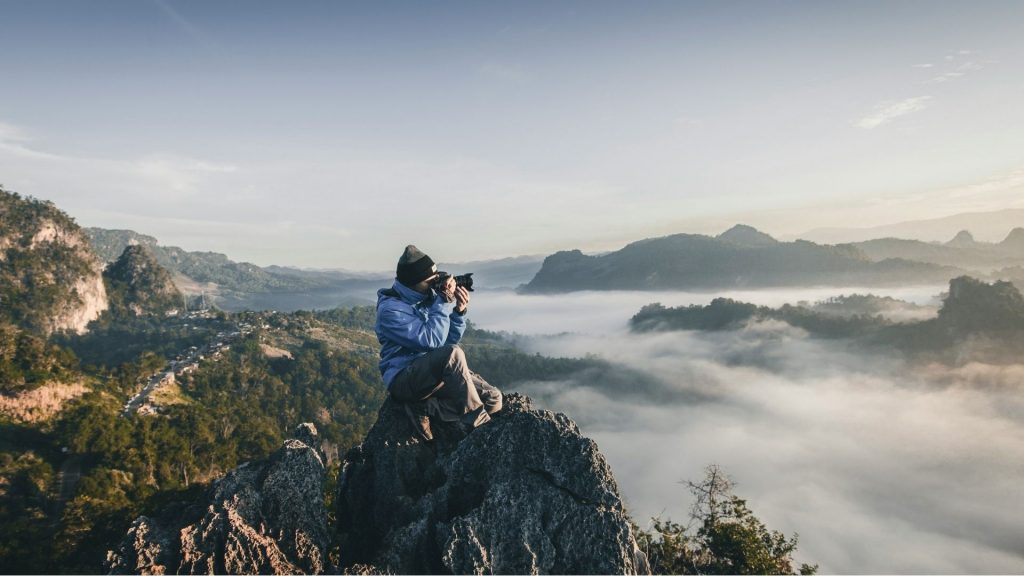 Photographer standing on a mountain above clouds capturing a landscape, representing why authenticity is winning in photography in the age of AI.