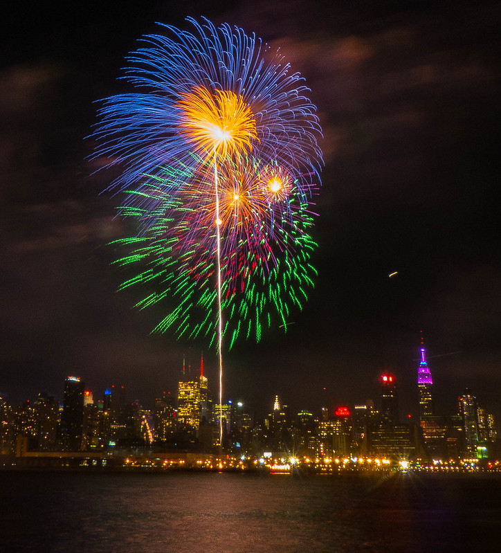 Fireworks Photography 101: long exposure of a blue, gold, and green firework burst rising over a city skyline with a vertical light trail reflected on the water
