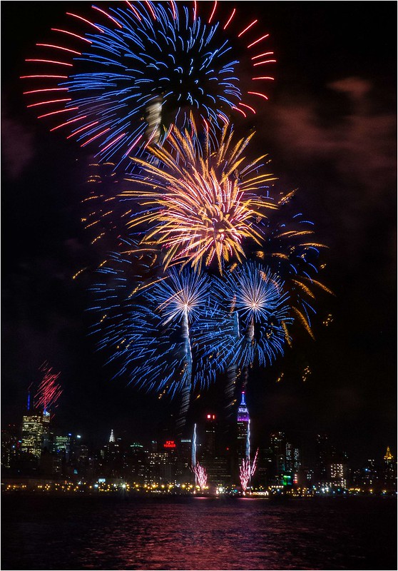 Fireworks Photography 101 example — blue, red, and gold fireworks over the New York City skyline reflected on the water at night