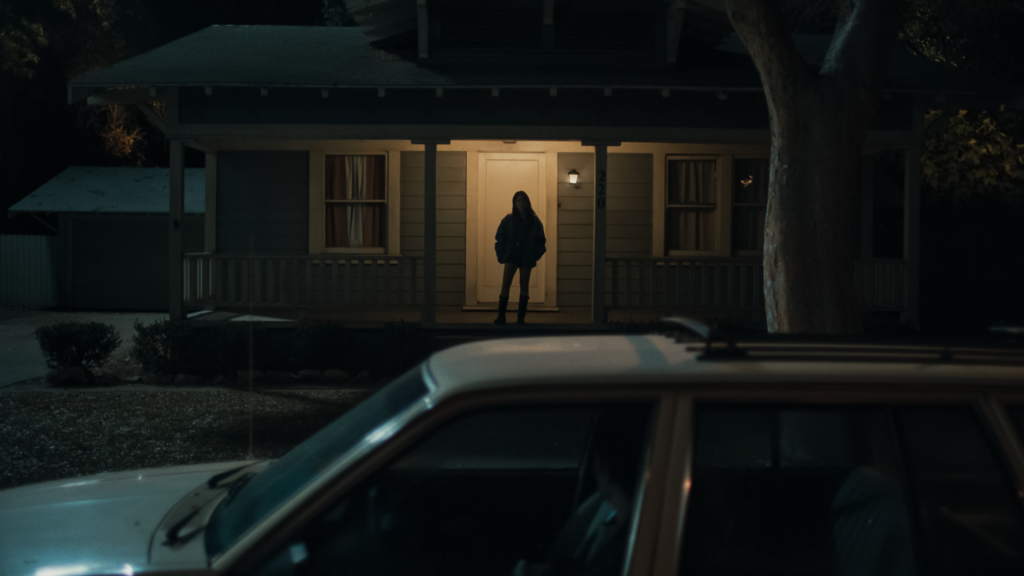 A dark, static wide shot from "Obsession": a lone silhouette in a lit doorway of a house at night.