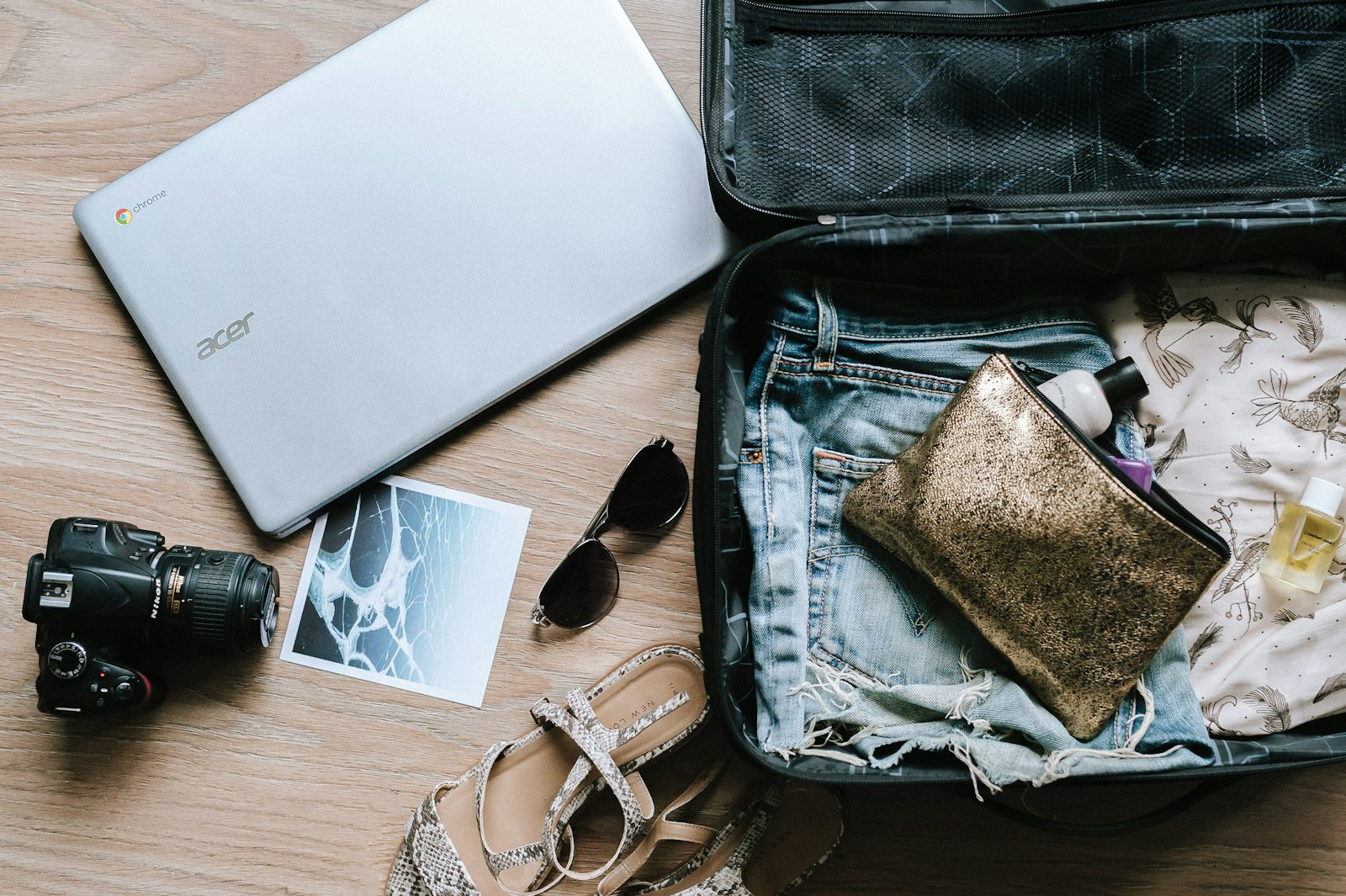 black DSLR camera beside a laptop, sunglasses, and an open suitcase on a wooden surface