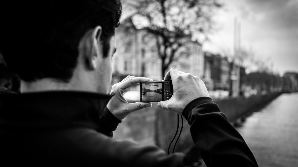 person holding a black compact camera photographing a city street and river in black and white