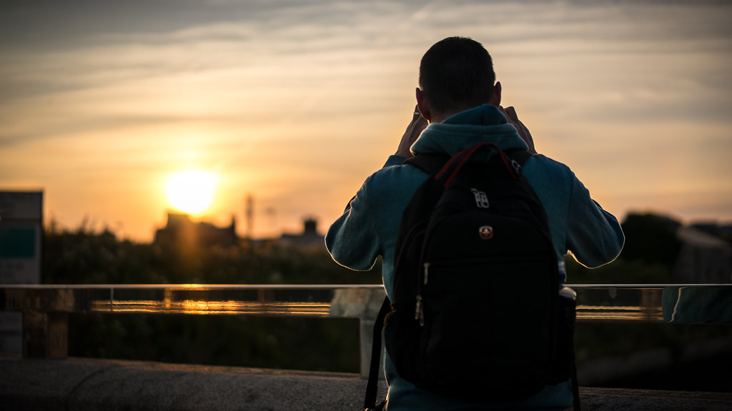 person with backpack photographing a city skyline at sunset from a railing
